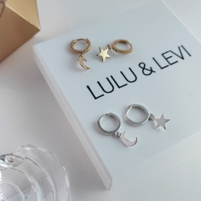 a pair of silver moon and star earrings and a gold pair of the same earrings laid out on white lulu and levi gift box with crystal and gold features in the background