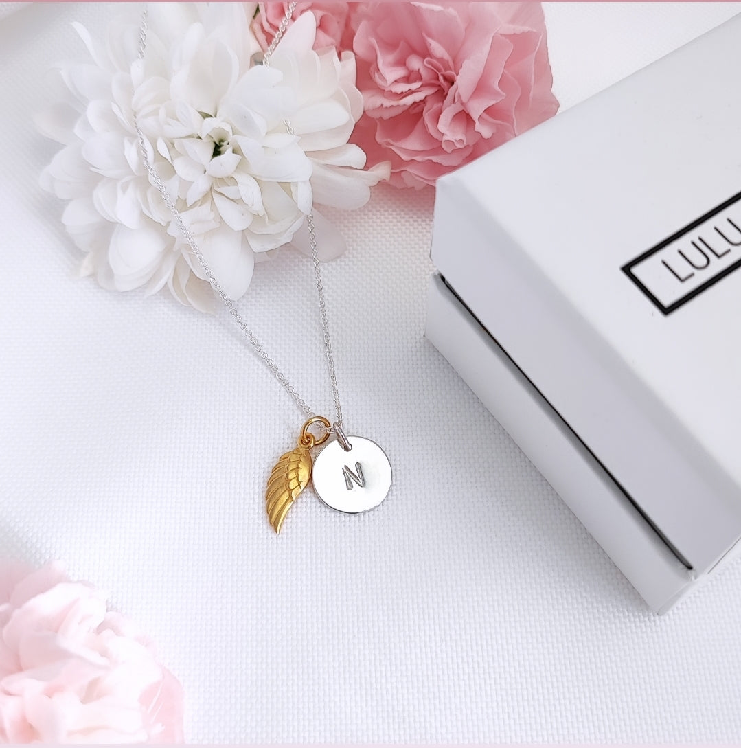 Silver personalised disc necklace with Gold Angel Wing on a white background with soft pink and white flowers beside a Lulu and Levi gift Box