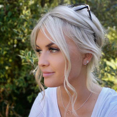 Blonde hair woman sitting outside wearing silver cirlce necklace and matching earrings and silver button earrings